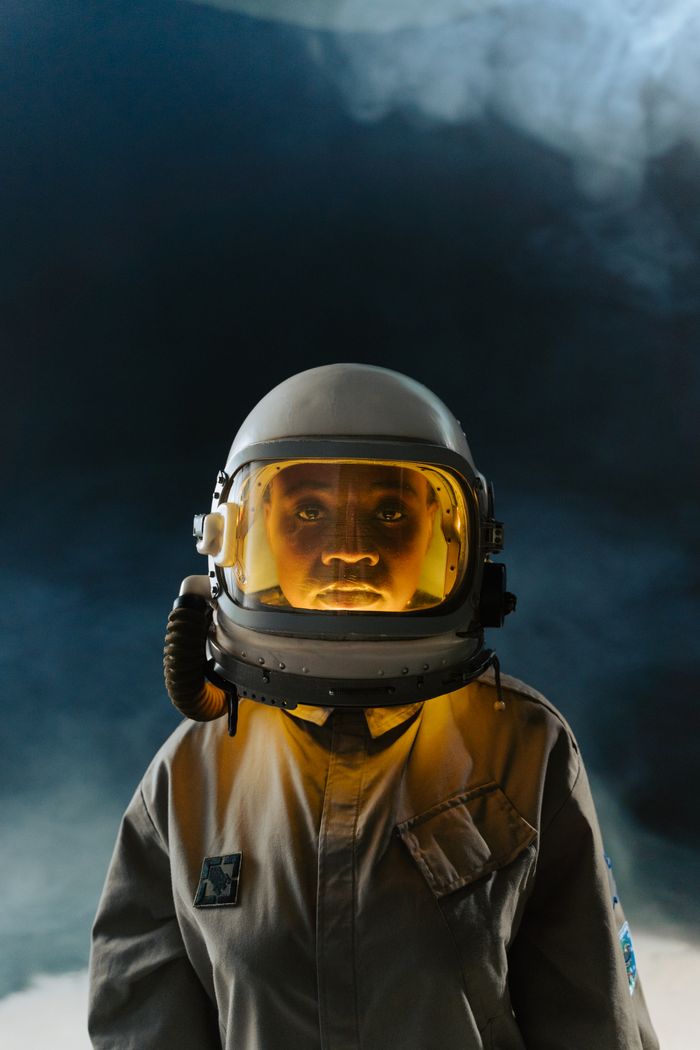 Portrait of an astronaut in a helmet, standing against a dark, cloud-filled backdrop, suggesting a cosmic or space theme.
