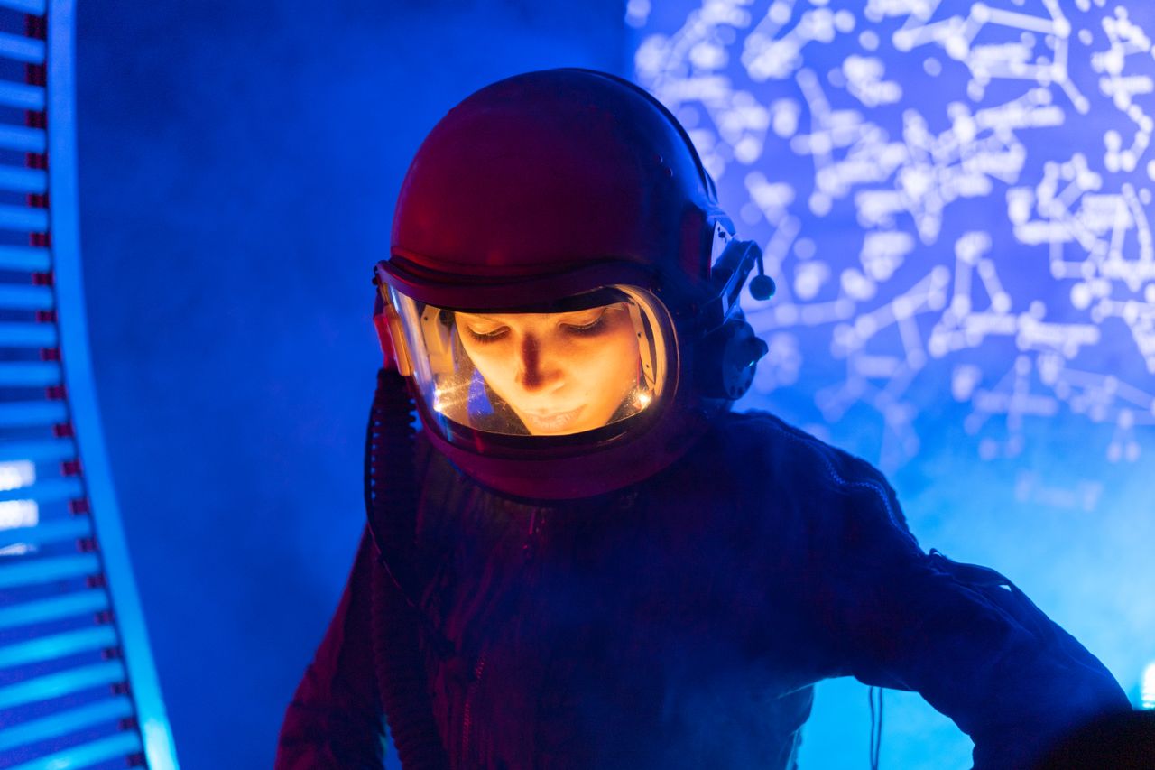 A woman in an astronaut suit looking down, with a glowing star map in the background, creating a futuristic space theme.