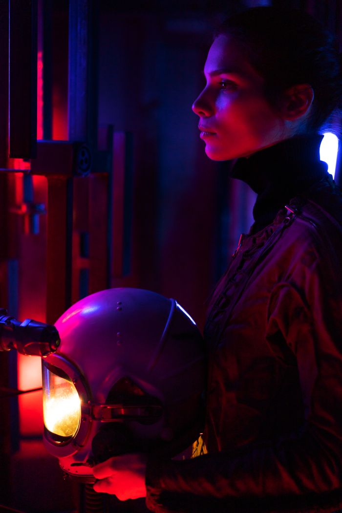 Profile of a spacewoman holding helmet, bathed in dramatic neon light.